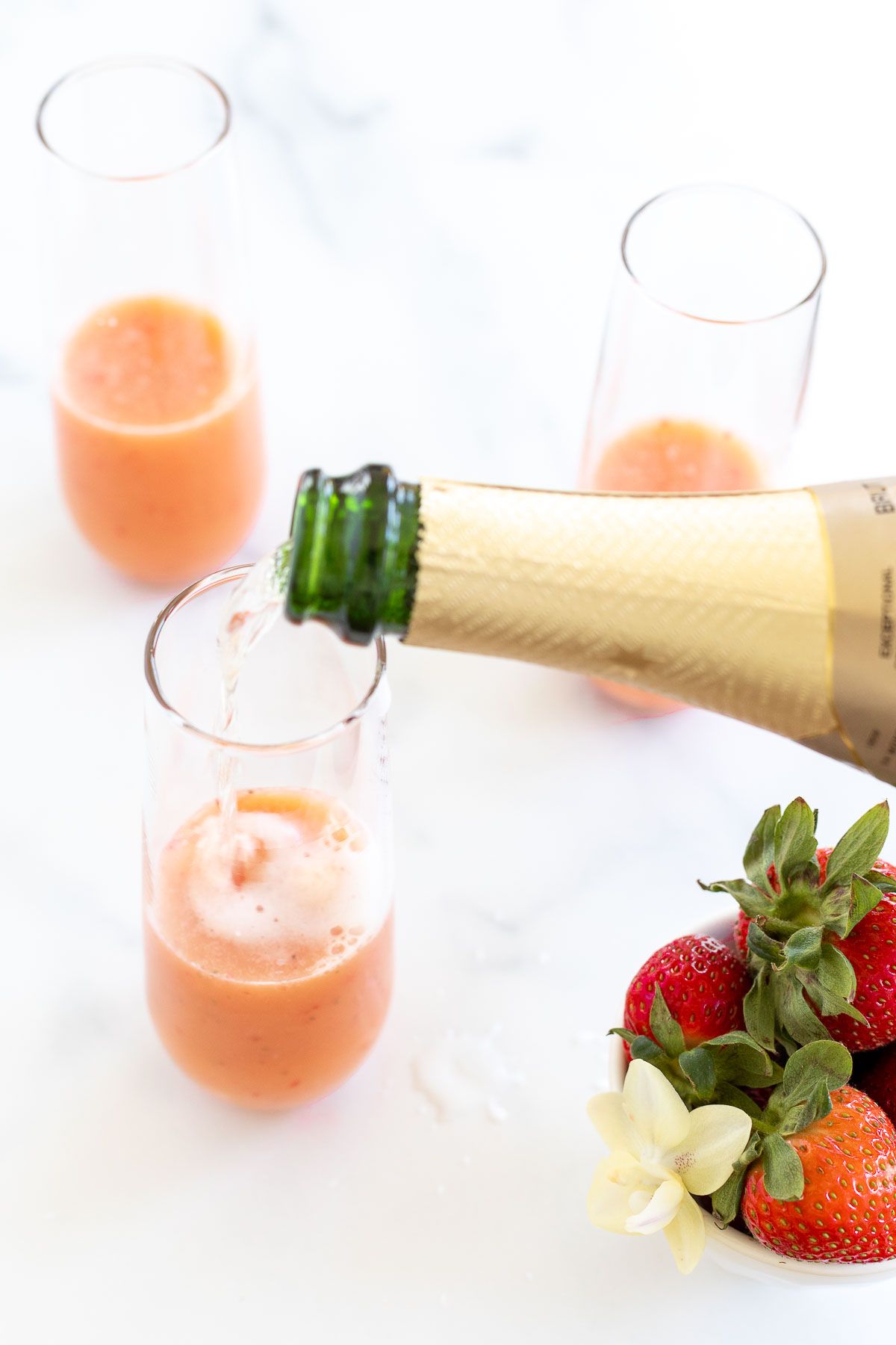 A bottle of Champagne pouring into strawberry mimosas, with a bowl of strawberries on the side