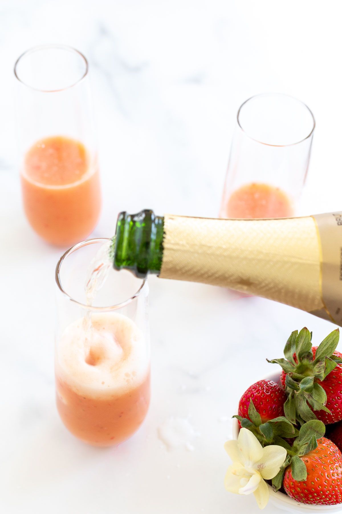 A bottle of Champagne pouring into strawberry mimosas, with a bowl of strawberries on the side