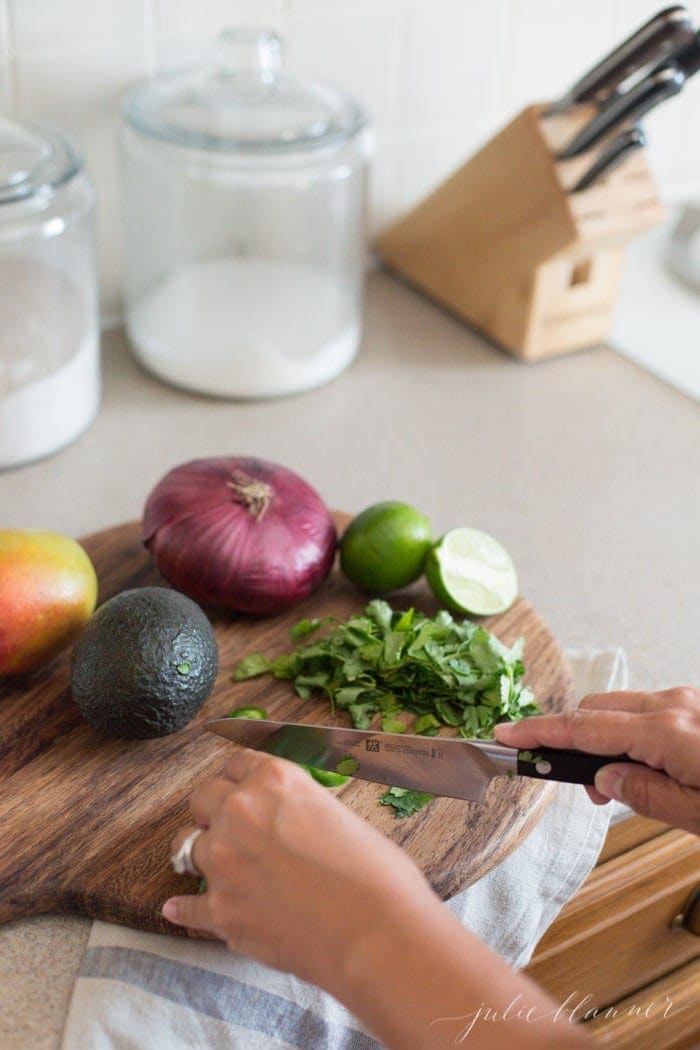 two hands slicing jalapeno with knife on wooden cutting board with avocado, onion, lime, cilantro