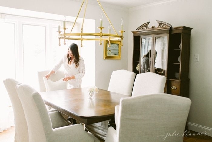 A brunette woman in a white dining room with  traditional furniture and a brass light fixture