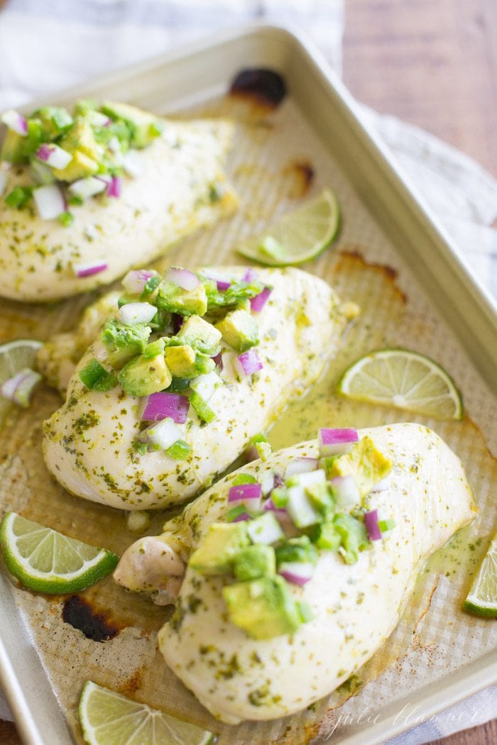 baked cilantro lime chicken topped with avocado salsa with lime wedges on baking sheet