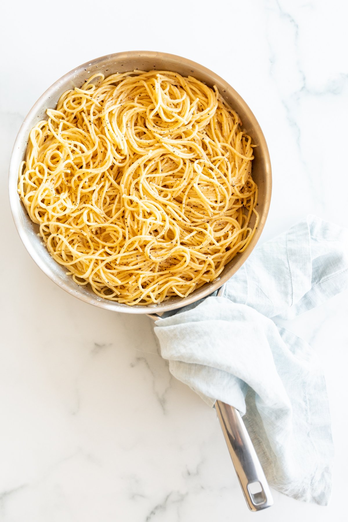 A silver pan full of cacio e pepe noodles, blue towel wrapped around handle on a marble surface.