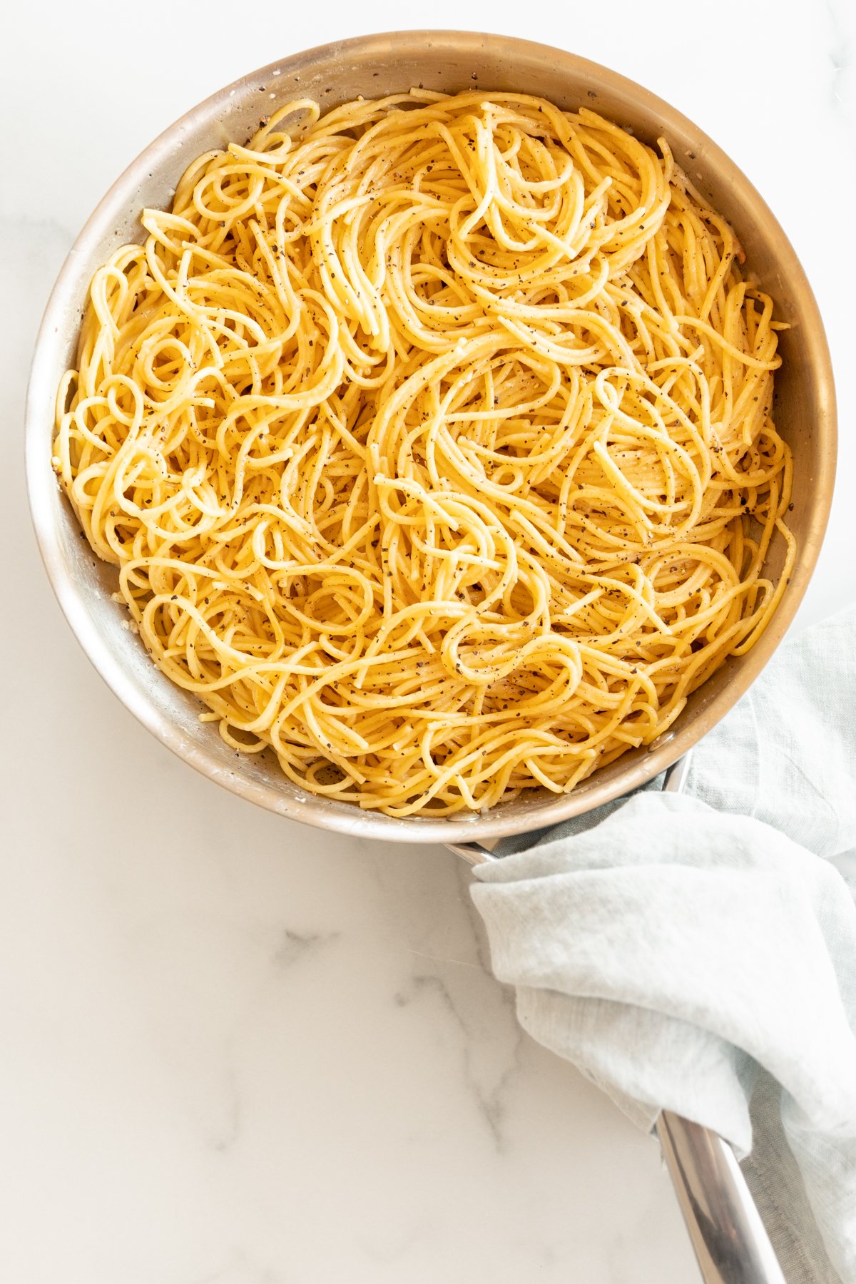 A silver pan full of cacio e pepe noodles, blue towel wrapped around handle on a marble surface.