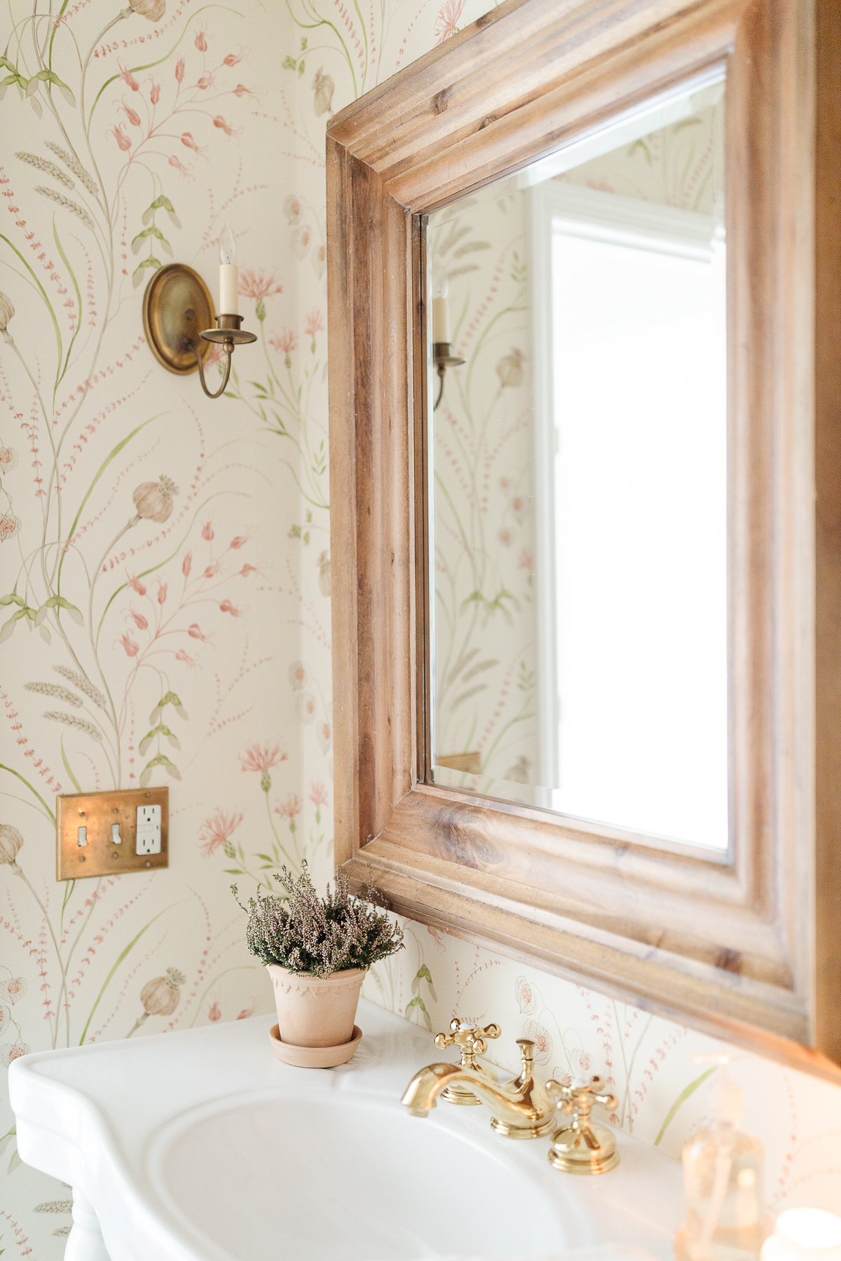 A wallpapered bathroom with a wood mirror and a brass switch plate cover