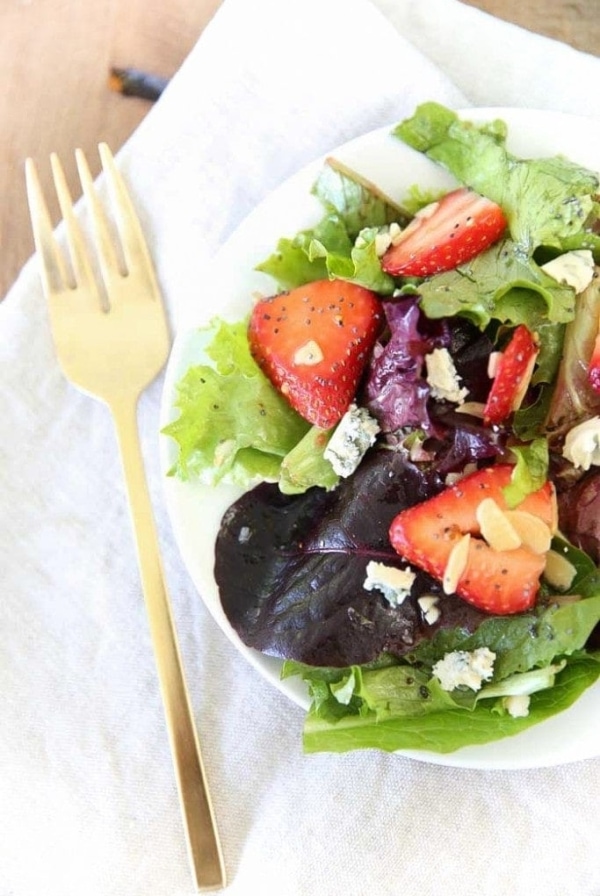 A white plate of salad covered in poppyseed dressing, gold fork to the side.