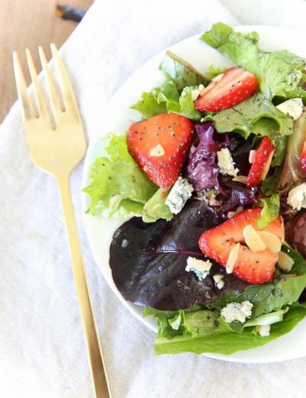 A white plate of salad covered in poppyseed dressing, gold fork to the side.