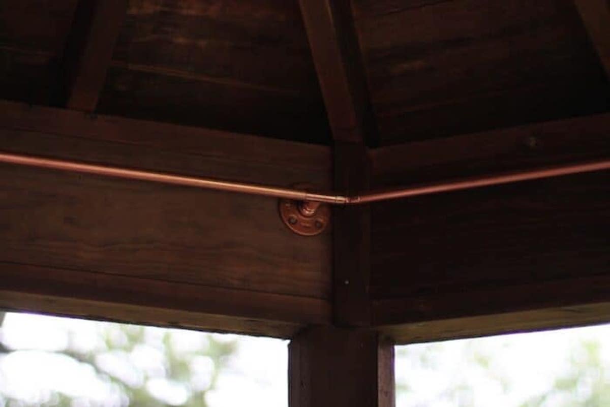 A copper pipe, which could be repurposed as a DIY copper curtain rod, is securely attached to a wooden ceiling frame with a bracket in this close-up view.