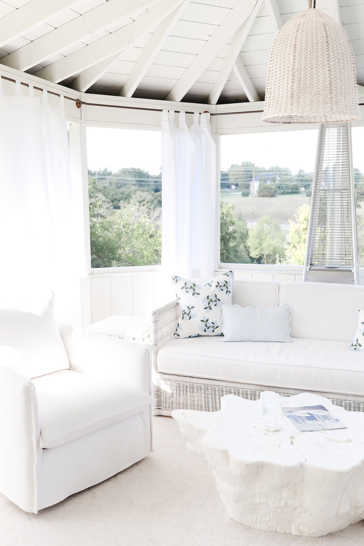 A bright, white sunroom with a cushioned sofa, armchair, tree-stump coffee table, hanging wicker light fixture, and large windows adorned with copper pipe curtain rods offering views of greenery outside.