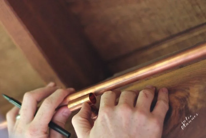 Hands holding and marking a copper pipe with a pen against a wooden surface, setting the stage for crafting DIY copper curtain rods.