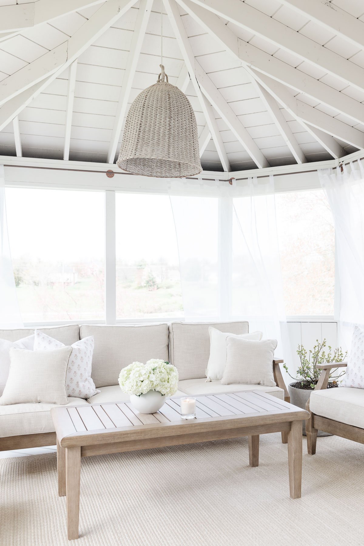 A bright, airy sunroom with white walls and ceiling, featuring a wooden table, cushioned seating with throw pillows, a hanging wicker light fixture, sheer curtains on DIY copper curtain rods, and a vase of white flowers on the table.