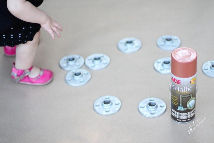 A toddler in pink shoes is near metallic piping discs arranged on a surface, with a can of Ace Metallic copper spray paint placed to the right, perfect materials for crafting DIY copper curtain rods.