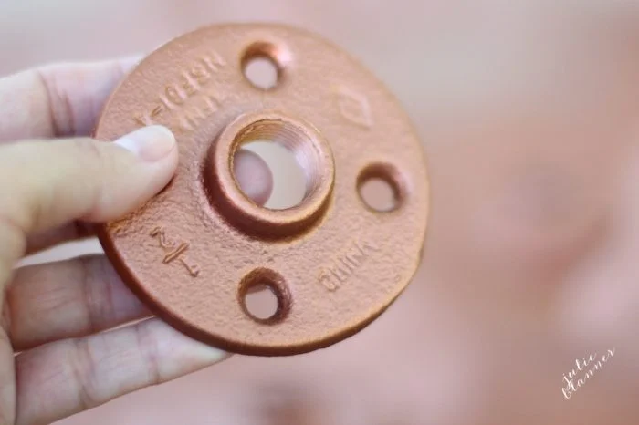 A hand holding a rust-colored metal flange with four holes and the word "China" engraved on it, perfect for DIY copper curtain rods.