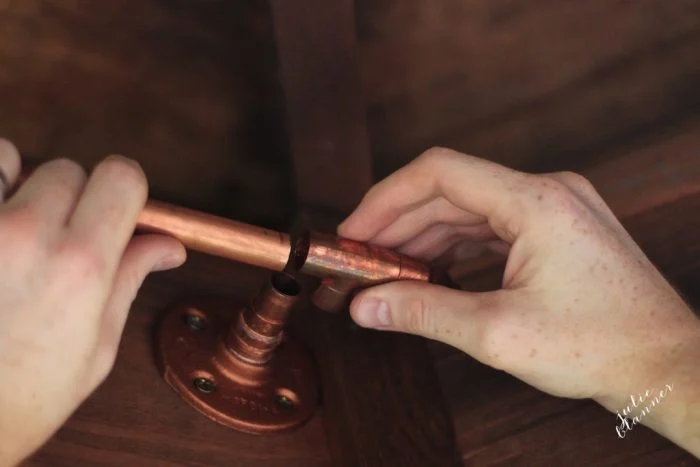 Two hands are shown repairing or connecting a copper plumbing pipe with a T-joint over a wooden surface, reminiscent of crafting DIY copper curtain rods.