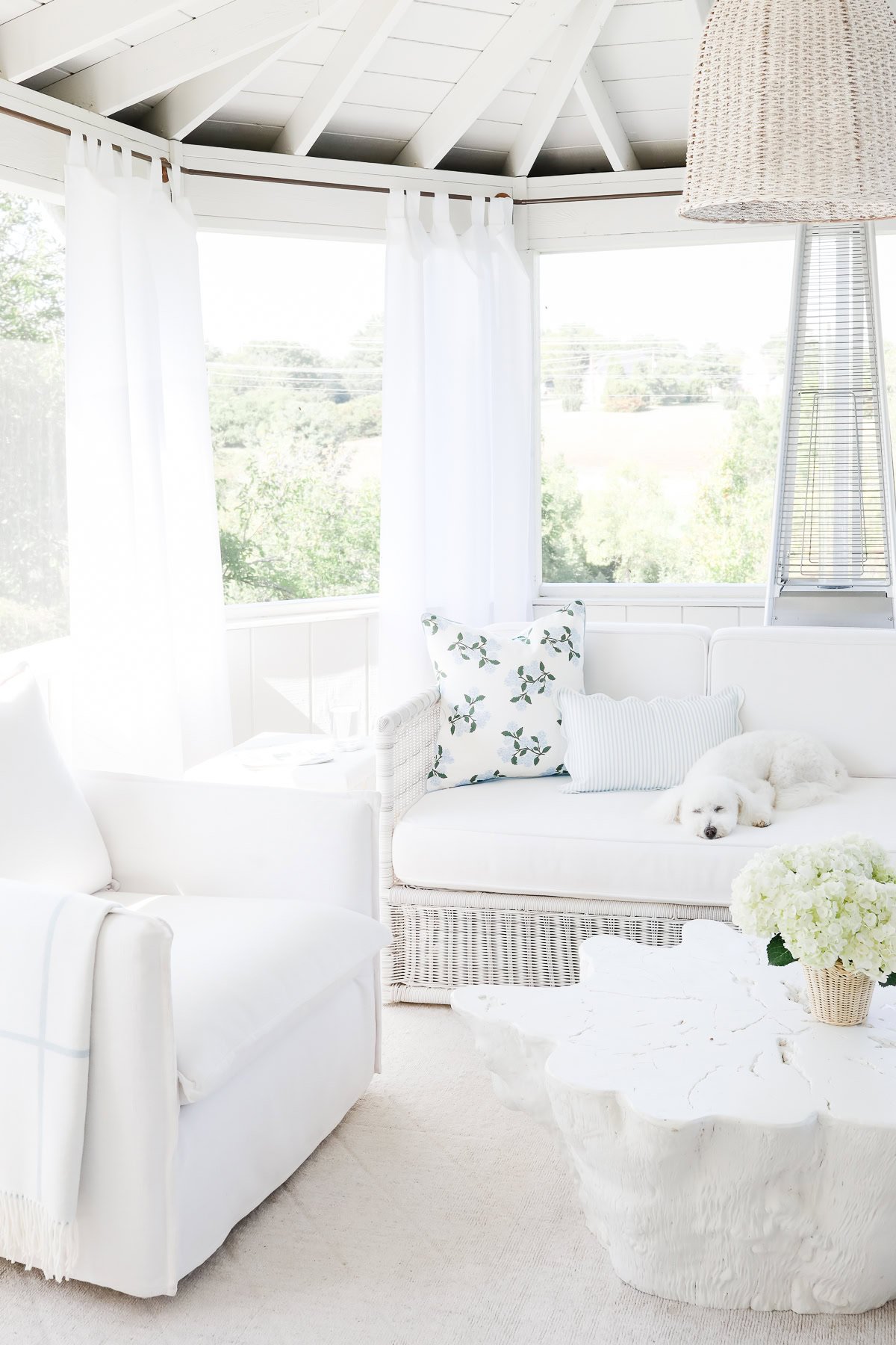 Bright sunroom with white furniture, a woven light fixture, a white dog on a sofa, floral pillows, a textured coffee table, and a vase of white hydrangeas. Large windows with copper pipe curtain rods let in natural light.
