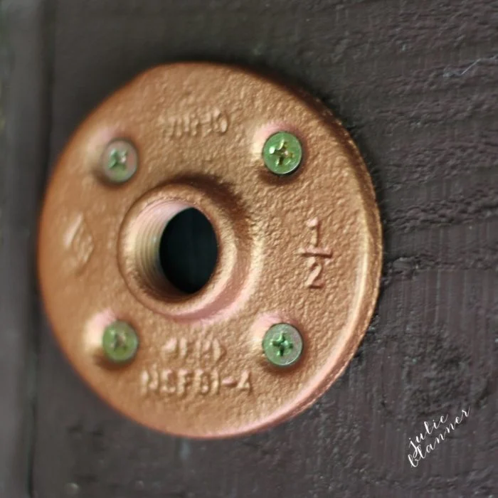 Close-up of a copper-colored round metal flange, reminiscent of diy copper curtain rods, attached to a dark surface with four green screws. The flange has engraved markings, including numbers and letters.
