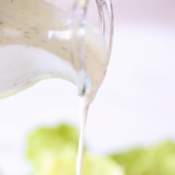 A clear glass bottle of homemade ranch dressing pouring over a salad