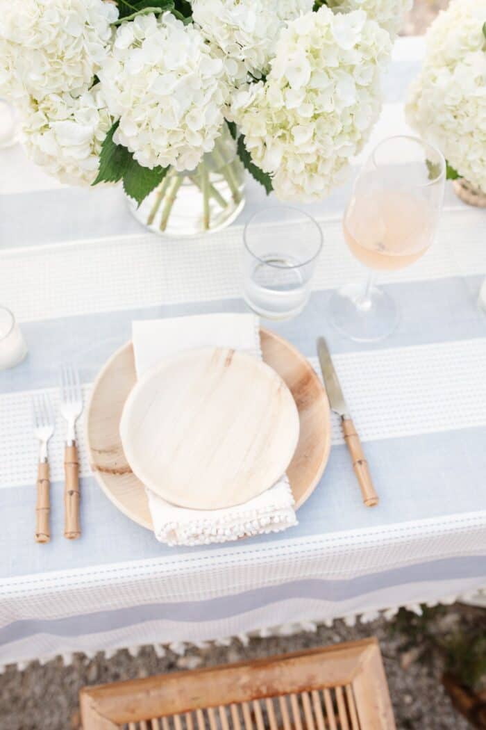 A wooden plate and charger with white flowers on a blue and white tablecloth for dinner al fresco.