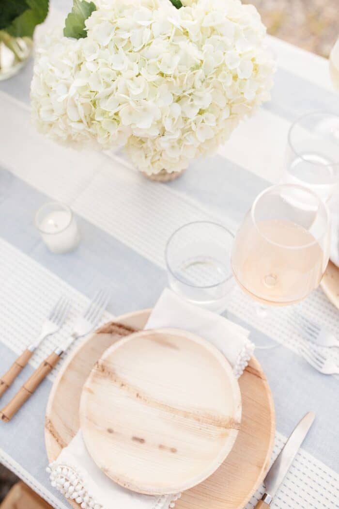 A wooden plate and charger with white flowers on a blue and white tablecloth for dinner al fresco.