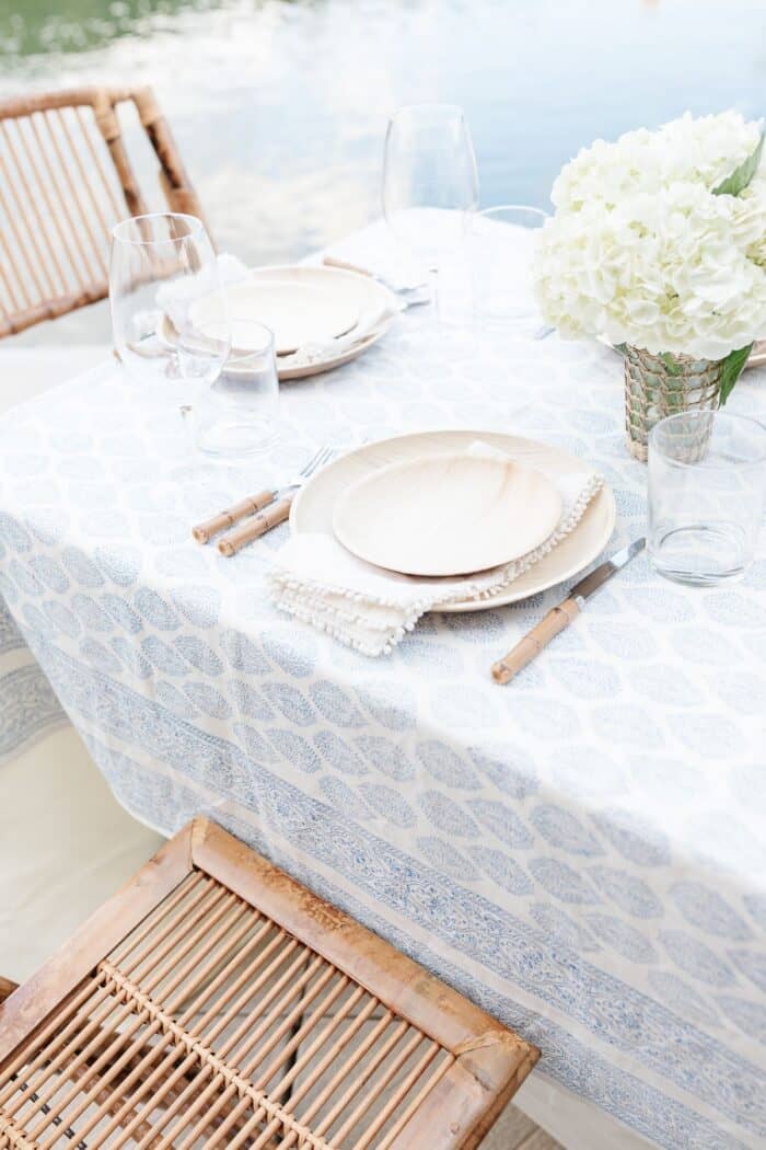An al fresco dining table set with white flowers and a blue and white table cloth, water in the background.