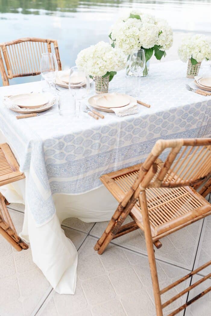 An al fresco dining table set with white flowers and a blue and white table cloth, water in the background.