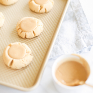 Peanut butter caramel cookies on a baking sheet with a bowl of peanut butter.