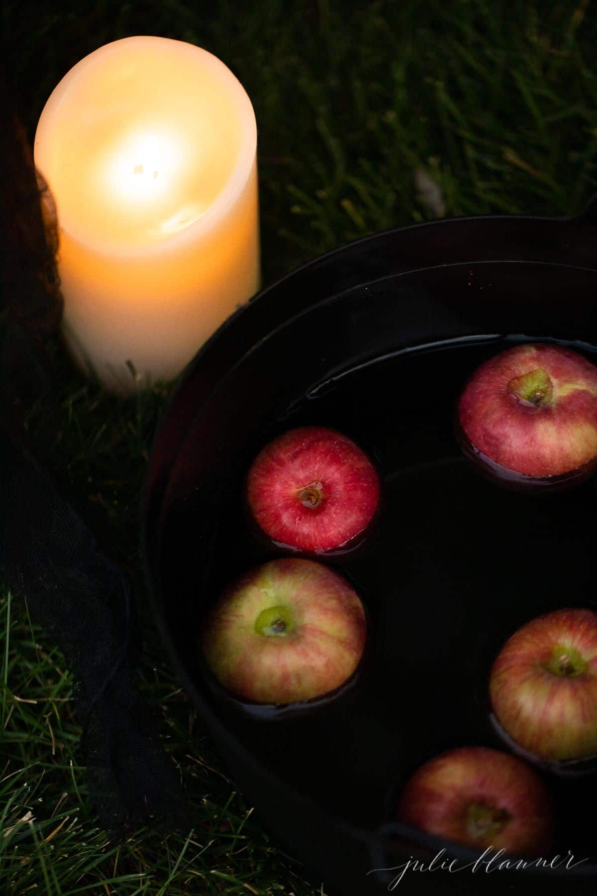 A cauldron full of fresh apples and a battery powered pillar candle sitting in grass.