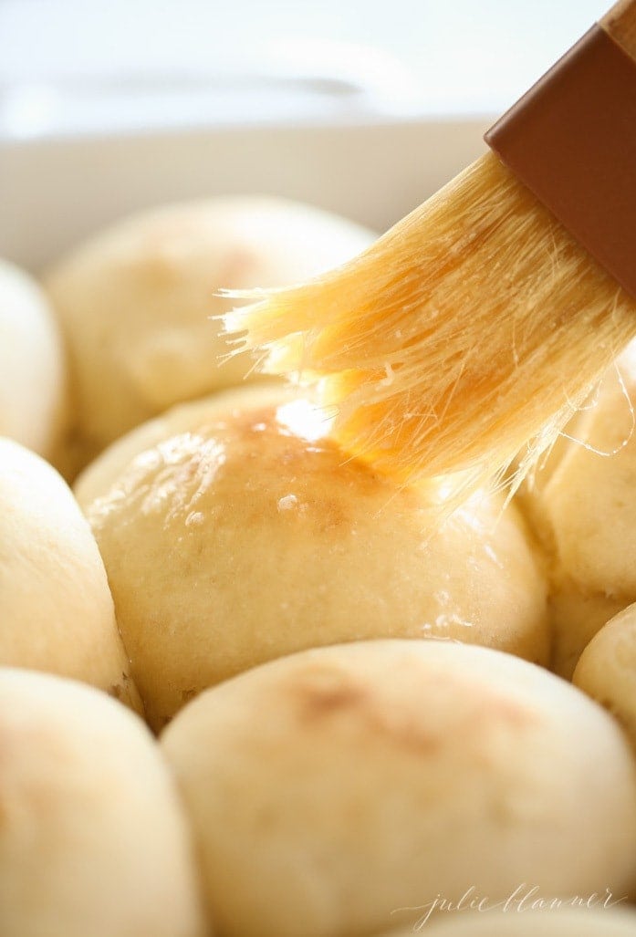 A pan full of homemade yeast rolls with a brush brushing melted butter on top.