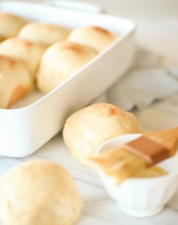 Dinner rolls in a white baking pant, with two in the foreground getting brushed with butter.