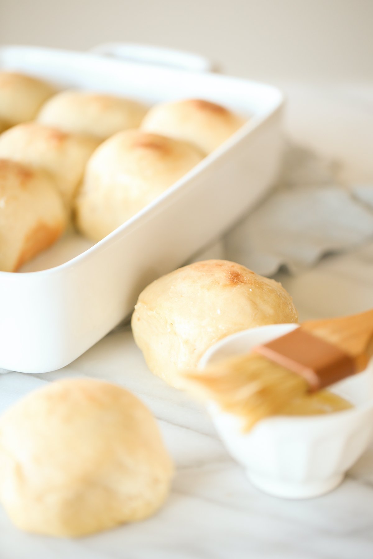 Dinner rolls in a white baking pant, with two in the foreground getting brushed with butter. 