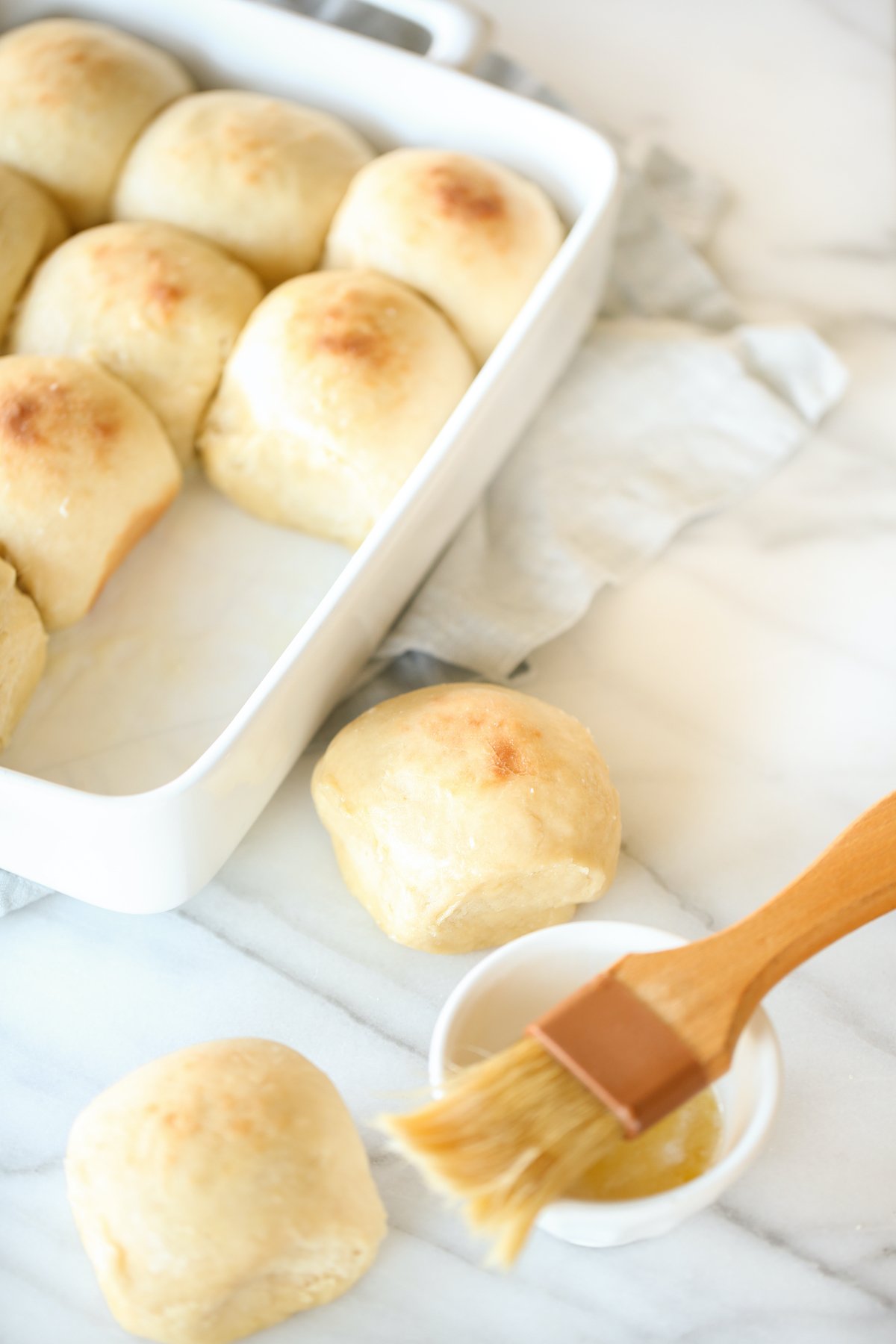 Dinner rolls in a white baking pant, with two in the foreground getting brushed with butter.