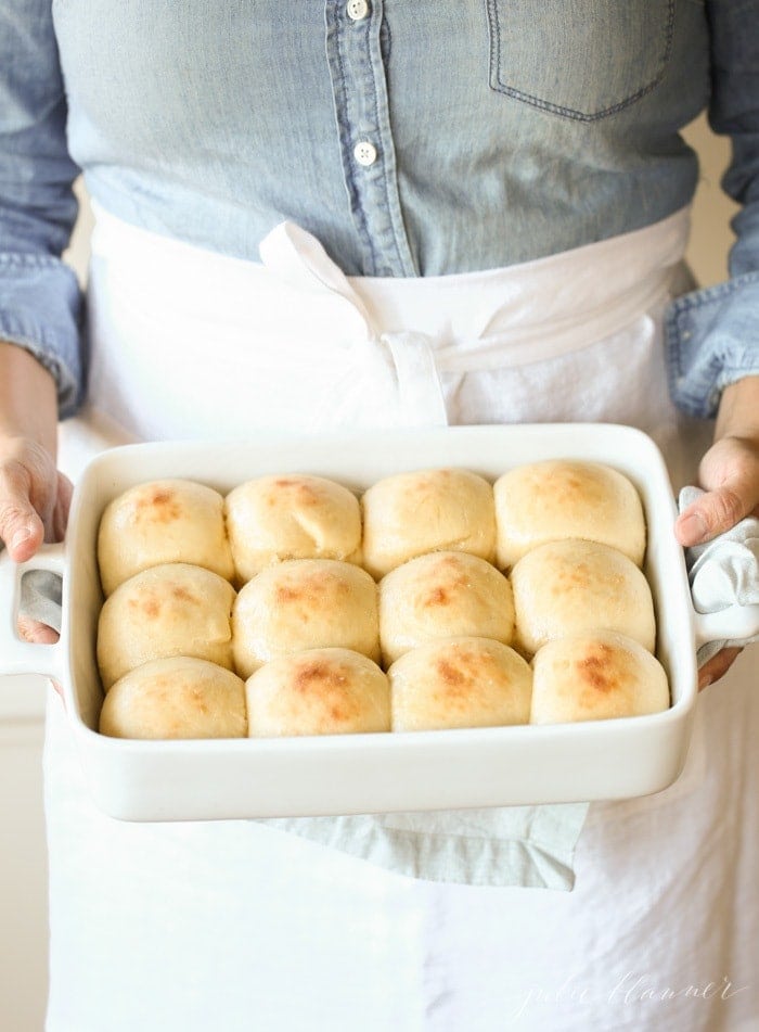 A woman in a blue shirt with a white apron at the waist, holding a white pan full of homemade dinner rolls.
