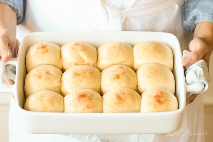 A woman in a blue shirt holding a white pan full of a homemade dinner roll recipe.
