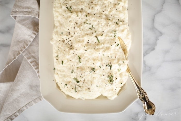 cauliflower mashed potatoes with rosemary in a white platter with gold spoon