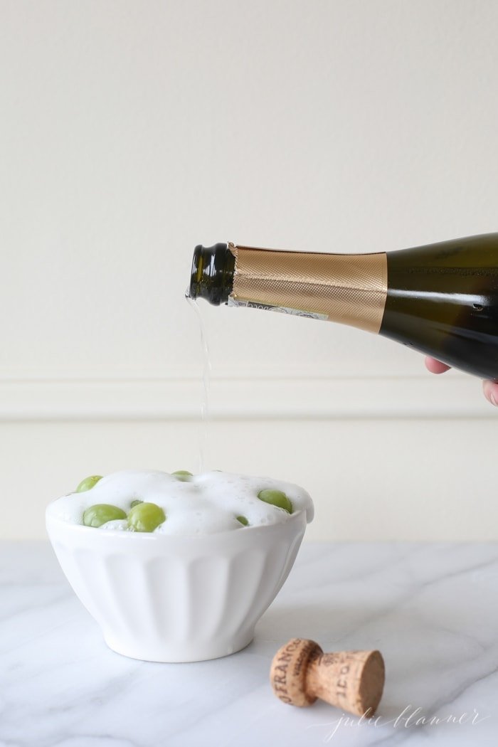 A bottle of champagne pouring over a bowl of green grapes.