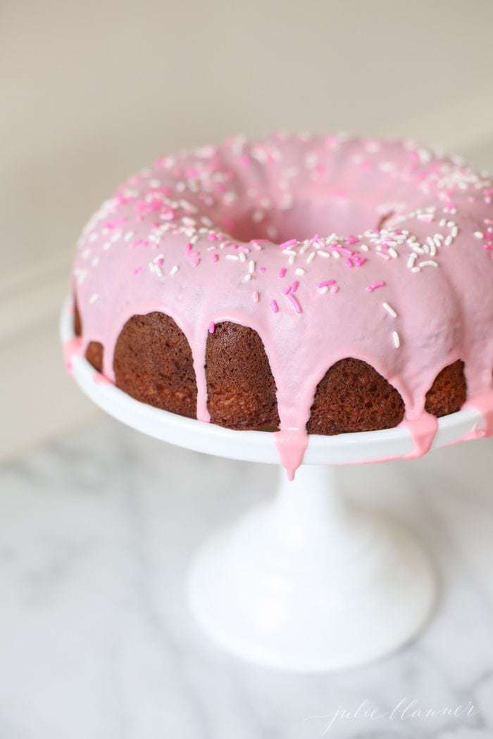 pink glazed donut cake with sprinkles on a white cake stand