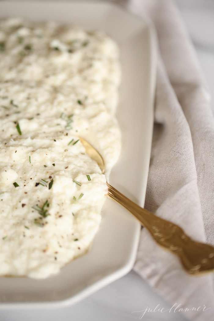 cauliflower mashed potatoes with rosemary is in a white platter with gold spoon