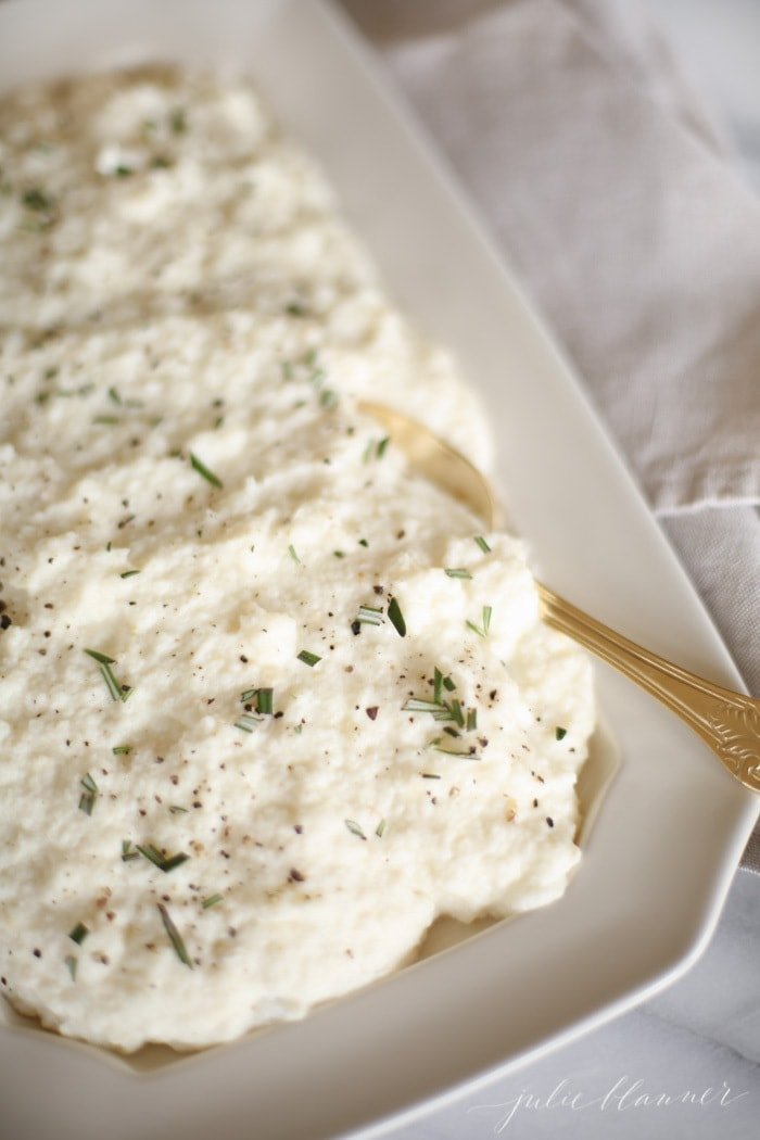 cauliflower mashed potatoes with rosemary in a white platter with gold spoon