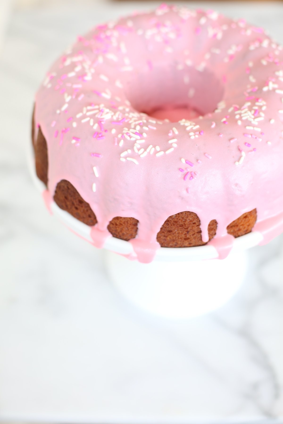 A donut cake on a white pedestal stand, decorated with pink frosting and sprinkles.