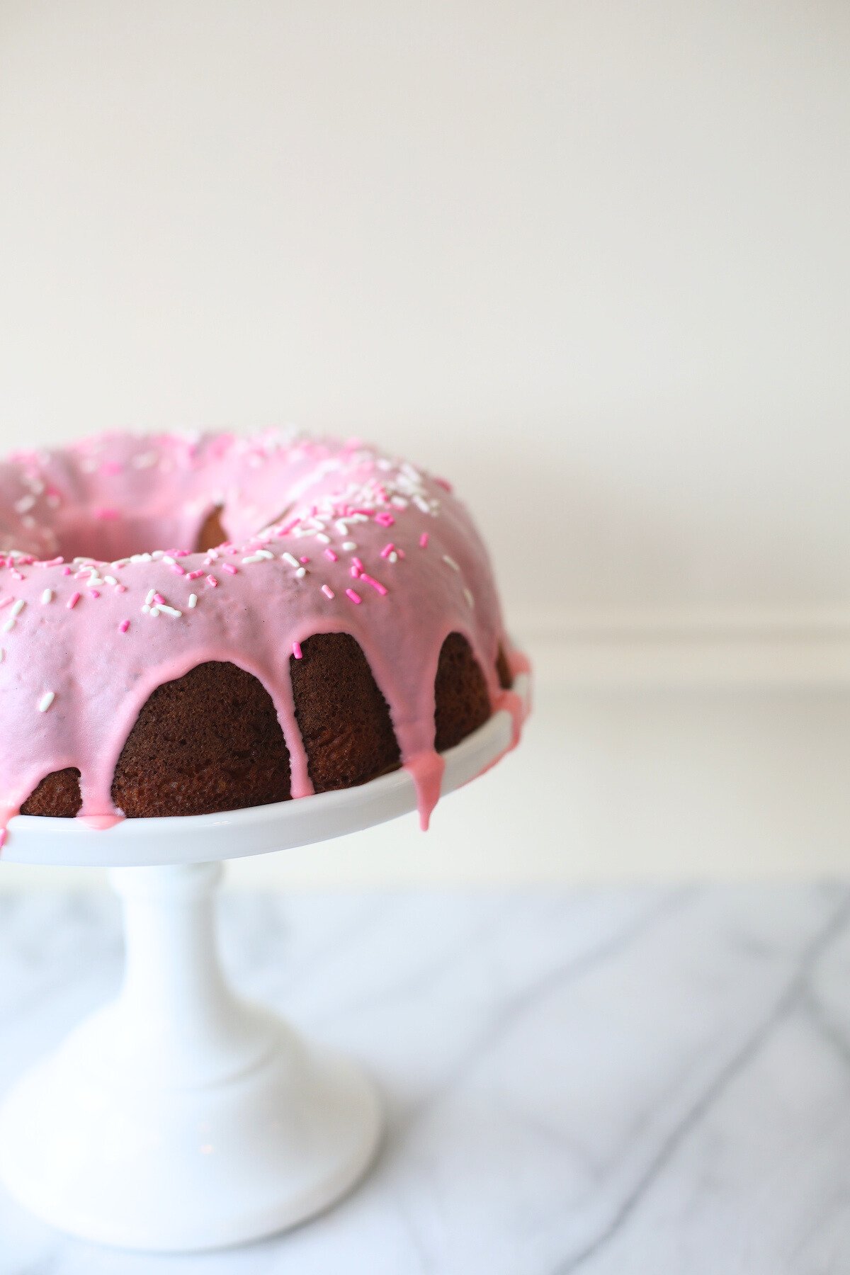 A donut cake on a white pedestal stand, decorated with pink frosting and sprinkles.