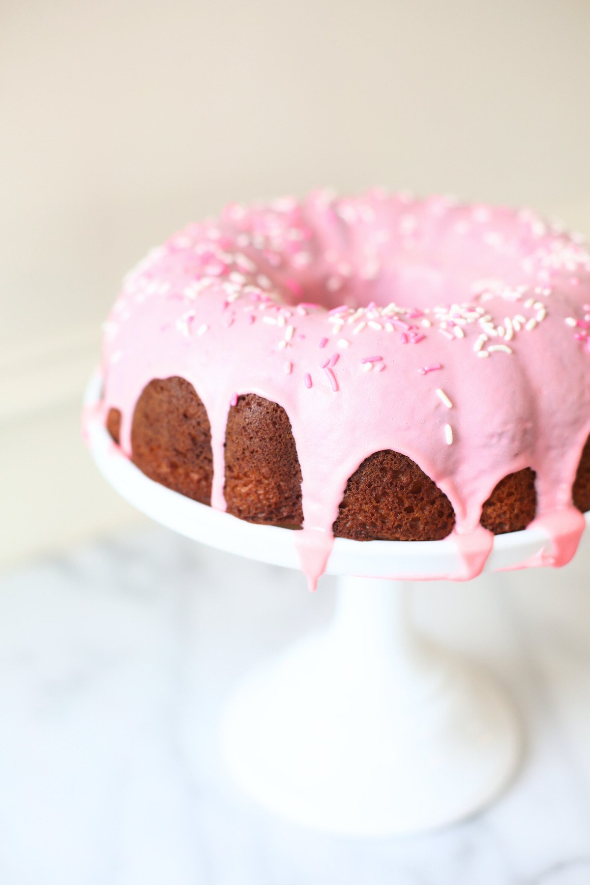 A donut cake on a white pedestal stand, decorated with pink frosting and sprinkles. 