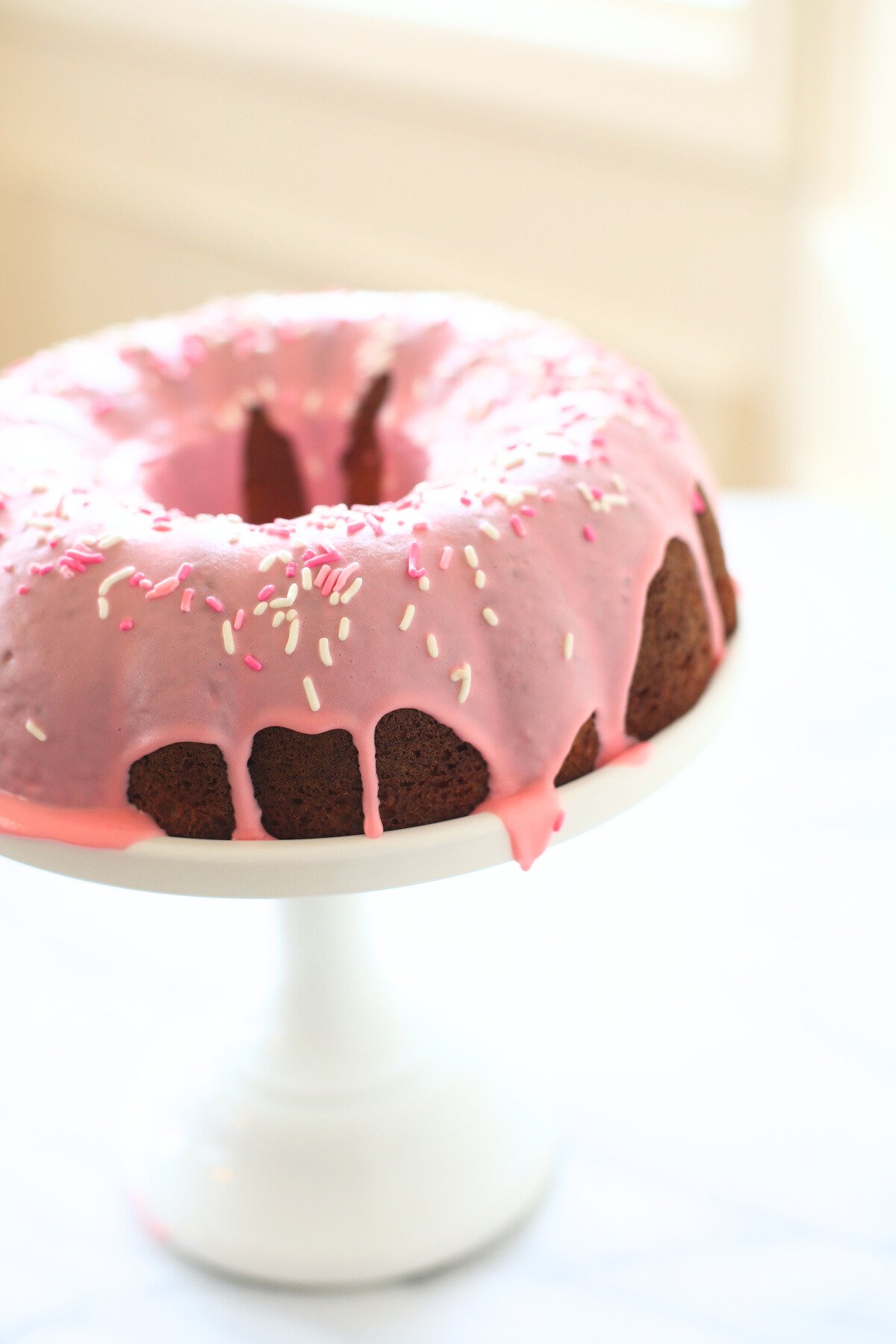 A donut cake on a white pedestal stand, decorated with pink frosting and sprinkles.