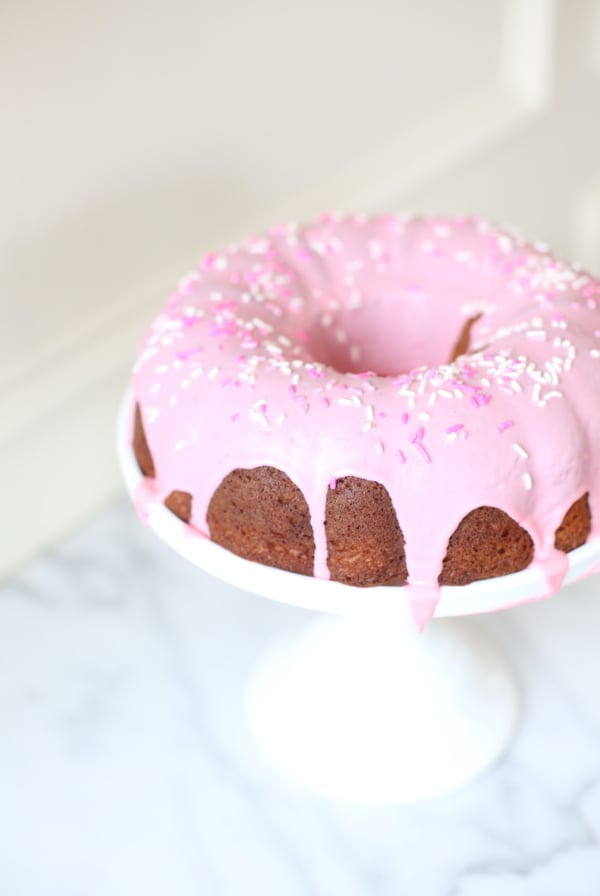 A donut cake on a white pedestal stand, decorated with pink frosting and sprinkles.