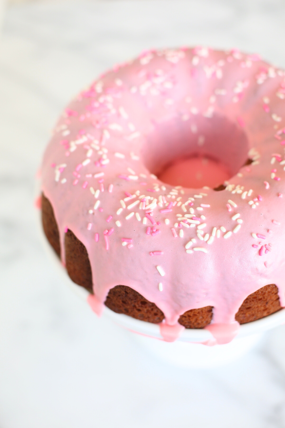 A donut cake on a white pedestal stand, decorated with pink frosting and sprinkles. 