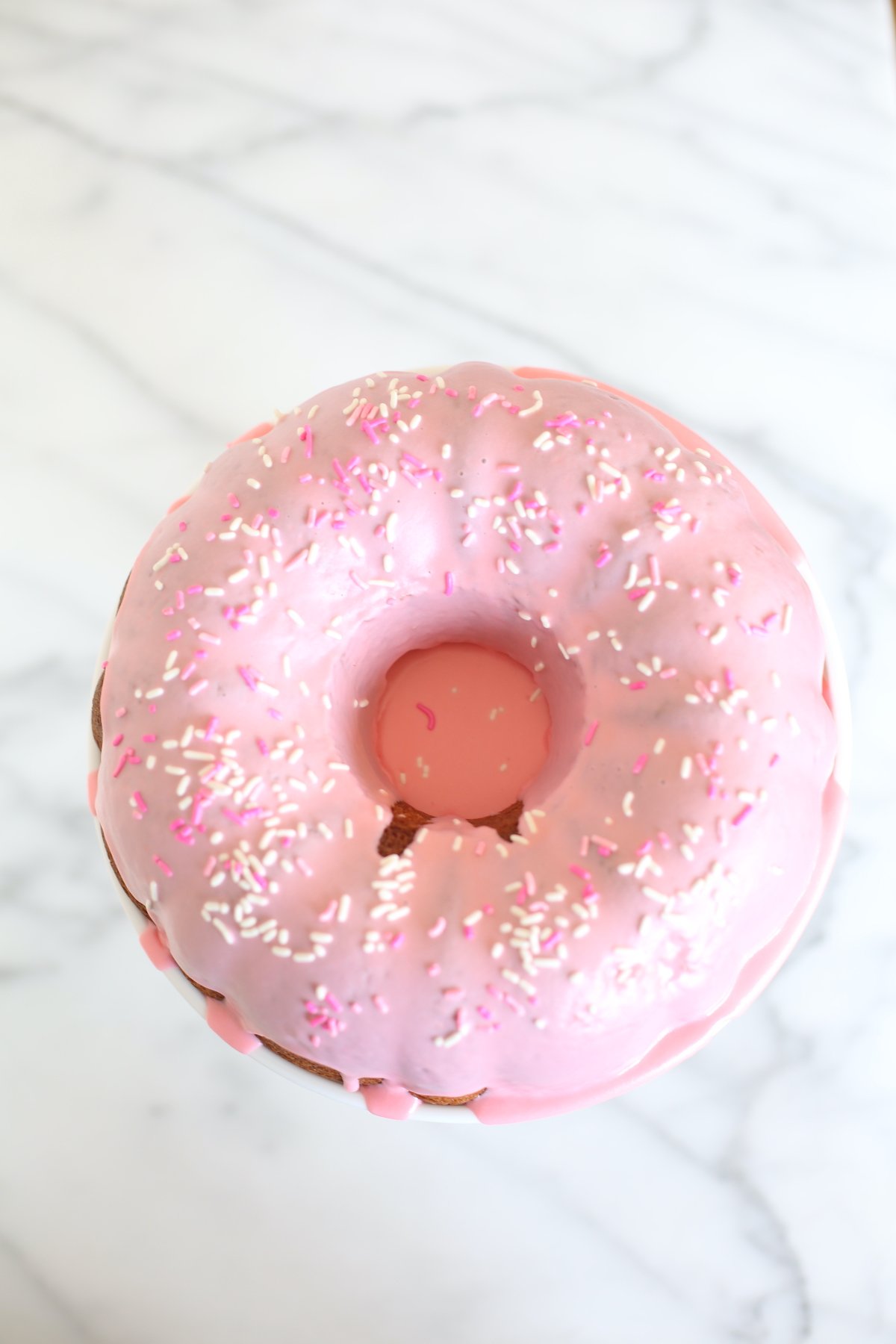 A donut cake on a white pedestal stand, decorated with pink frosting and sprinkles.