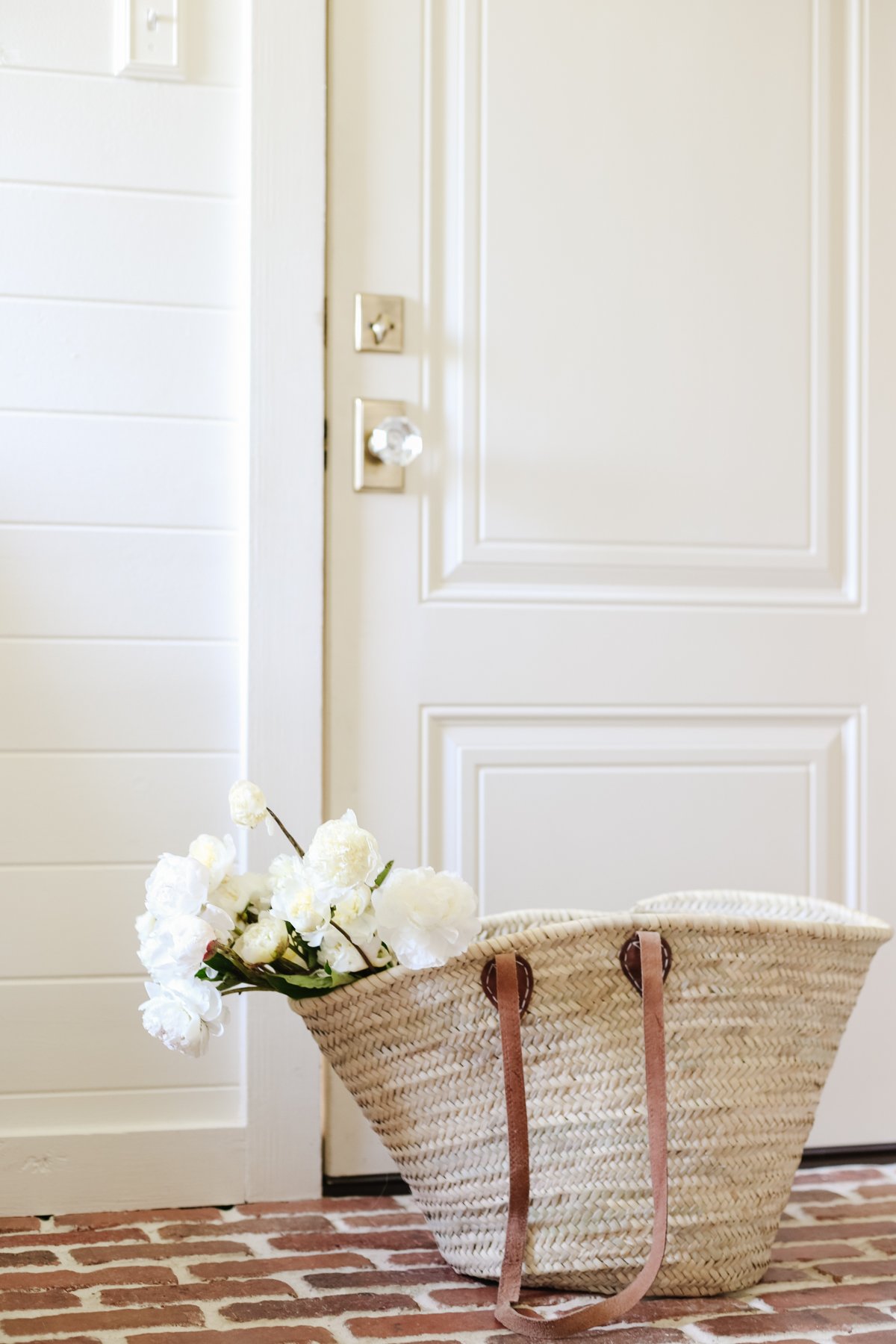 A raffia tote bag full of flowers on red brick flooring.