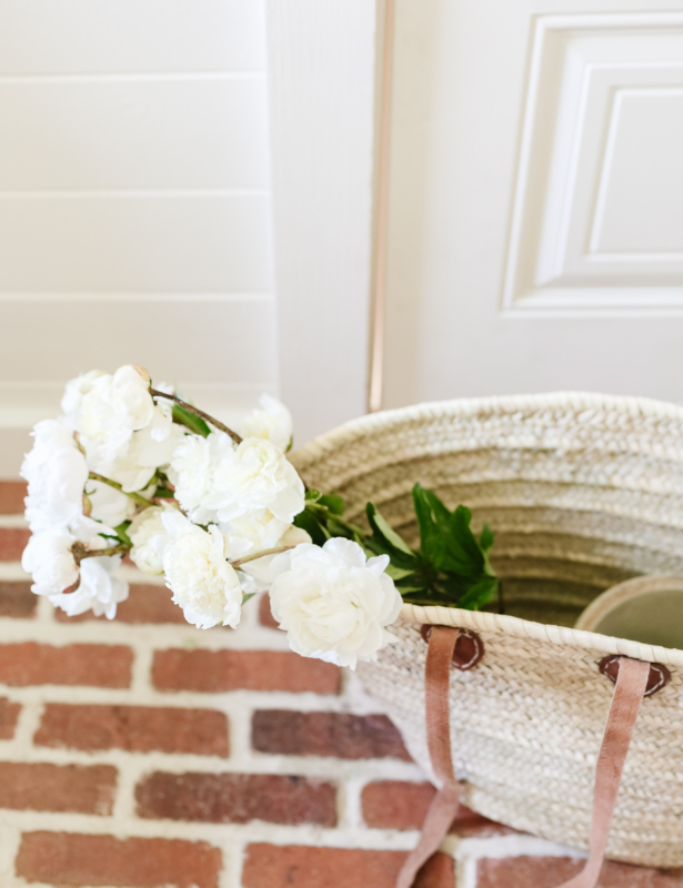 A raffia tote bag full of flowers on red brick flooring.