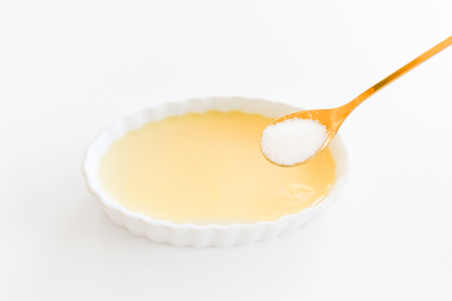 A spoonful of sugar sprinkled over a creamy creme brulee in a shallow white bowl on a white background.