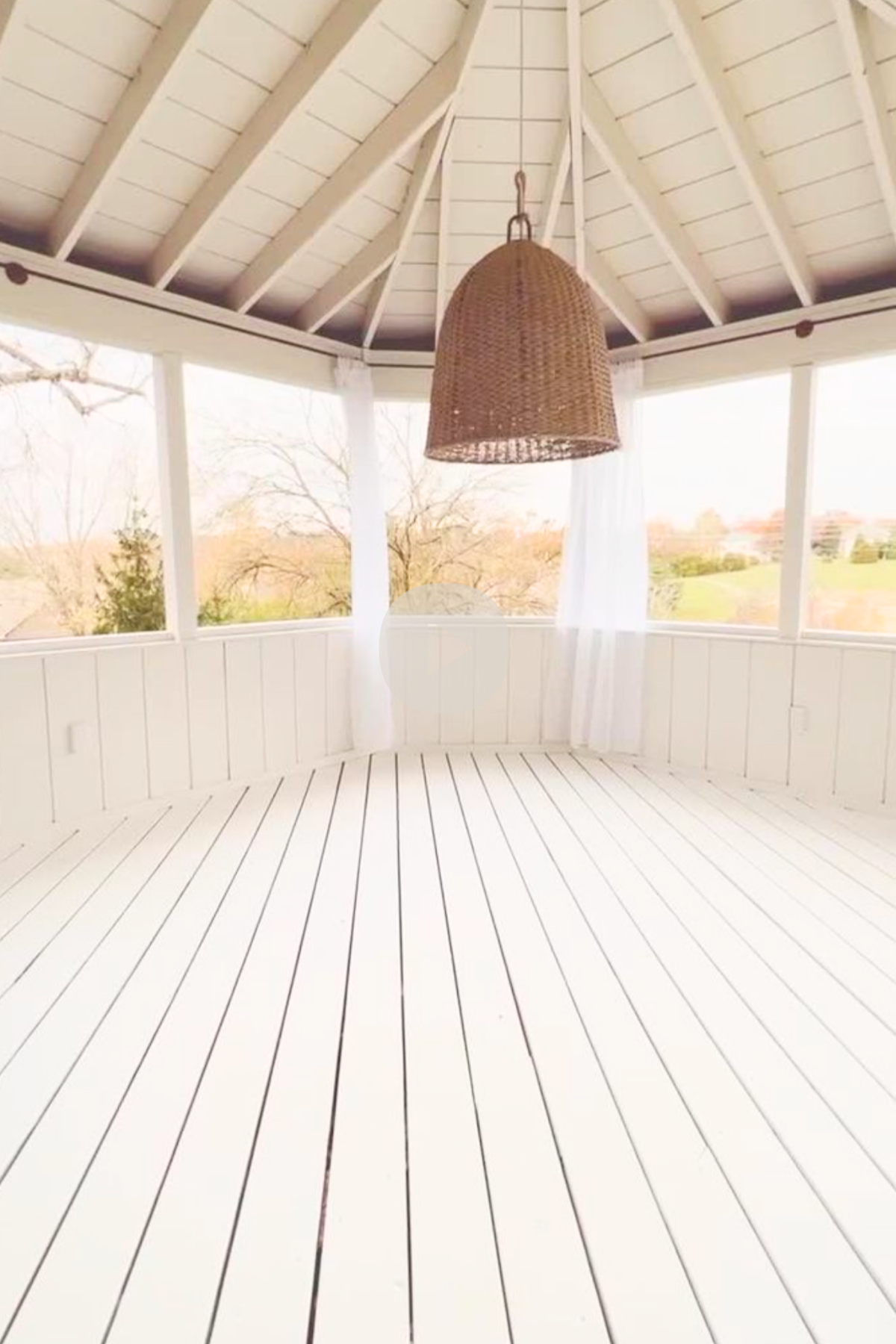 A white, wooden gazebo with a wicker hanging light fixture, open windows, and white floorboards that gleam as if treated with the best deck paint, overlooking a grassy landscape.