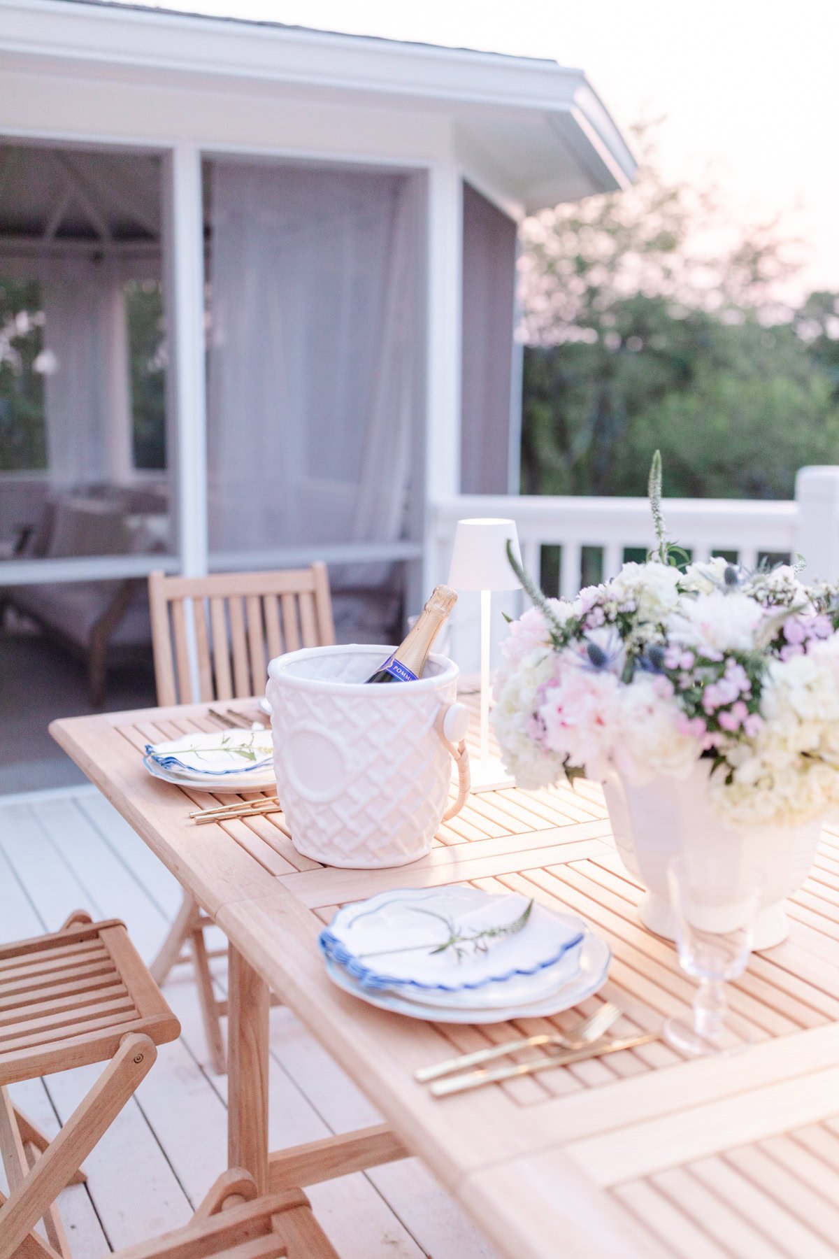 A beautifully stained wooden outdoor table set with plates, cutlery, a white flower arrangement, and a white ice bucket holding a bottle. Wooden chairs, also treated with the best deck stain for durability, are placed around the table. A charming house is in the background.