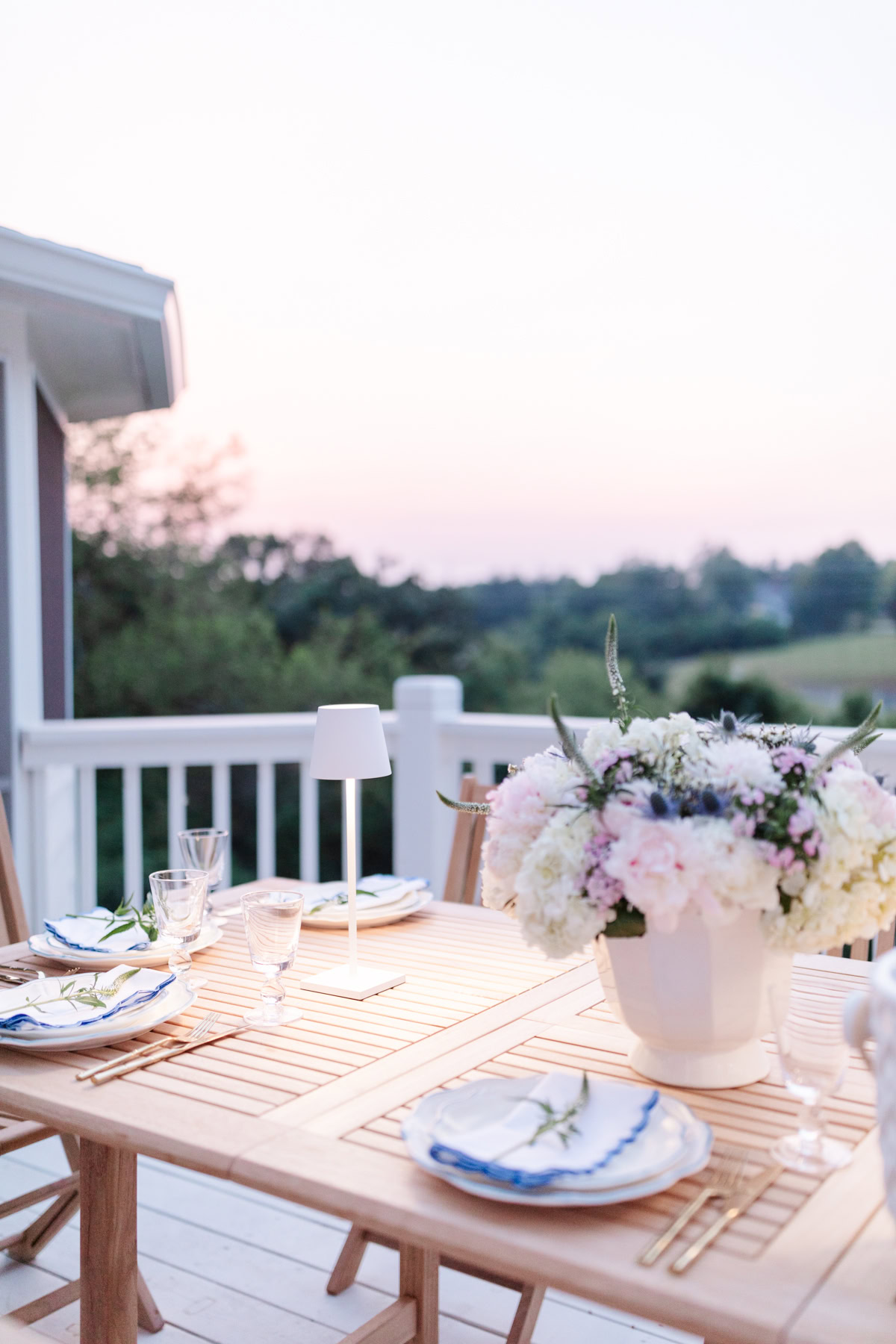 An outdoor dining table set with plates, glassware, and a floral centerpiece against a backdrop of trees and a pastel sky at sunset, showcasing the best deck stain to enhance your serene setting.